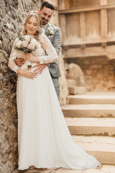 Hochzeitsfotograf Ingelheim Ein Brautpaar posiert verliebt vor einer steinernen Wand mit Blüten in der Hand.