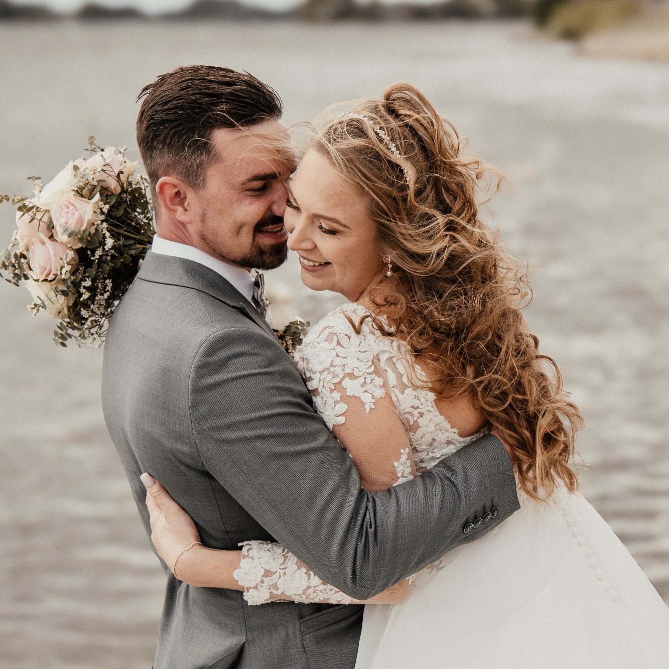 Hochzeitsfotograf Ingelheim - Brautpaar umarmt sich am Strand, lächelt und hält einen Blumenstrauß.
