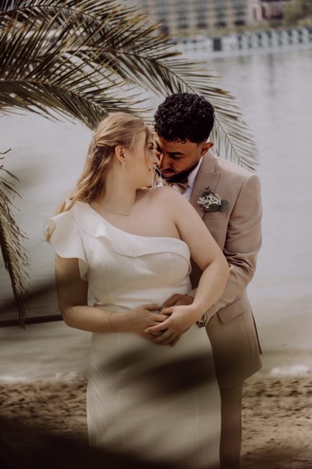 Hochzeitsfotograf Ingelheim Brautpaar umarmt sich am Strand, umgeben von Palmen und Wasser im Hintergrund.